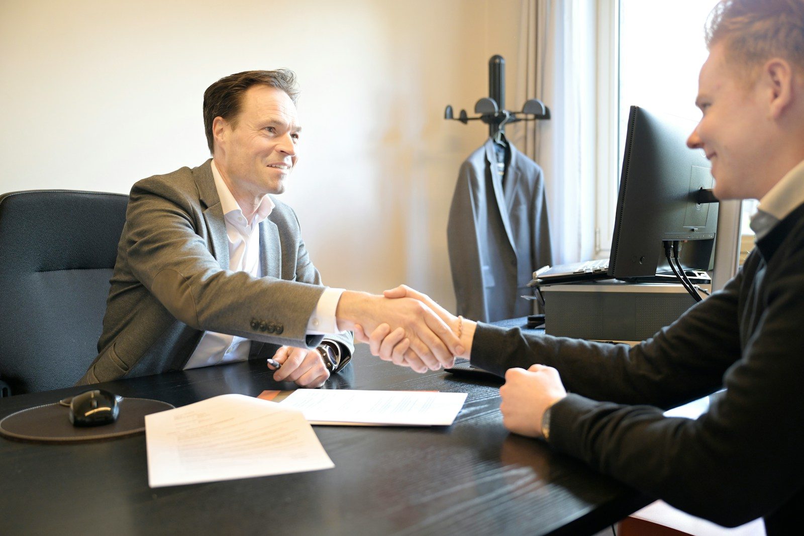 Two smiling men shake hands across an office desk.